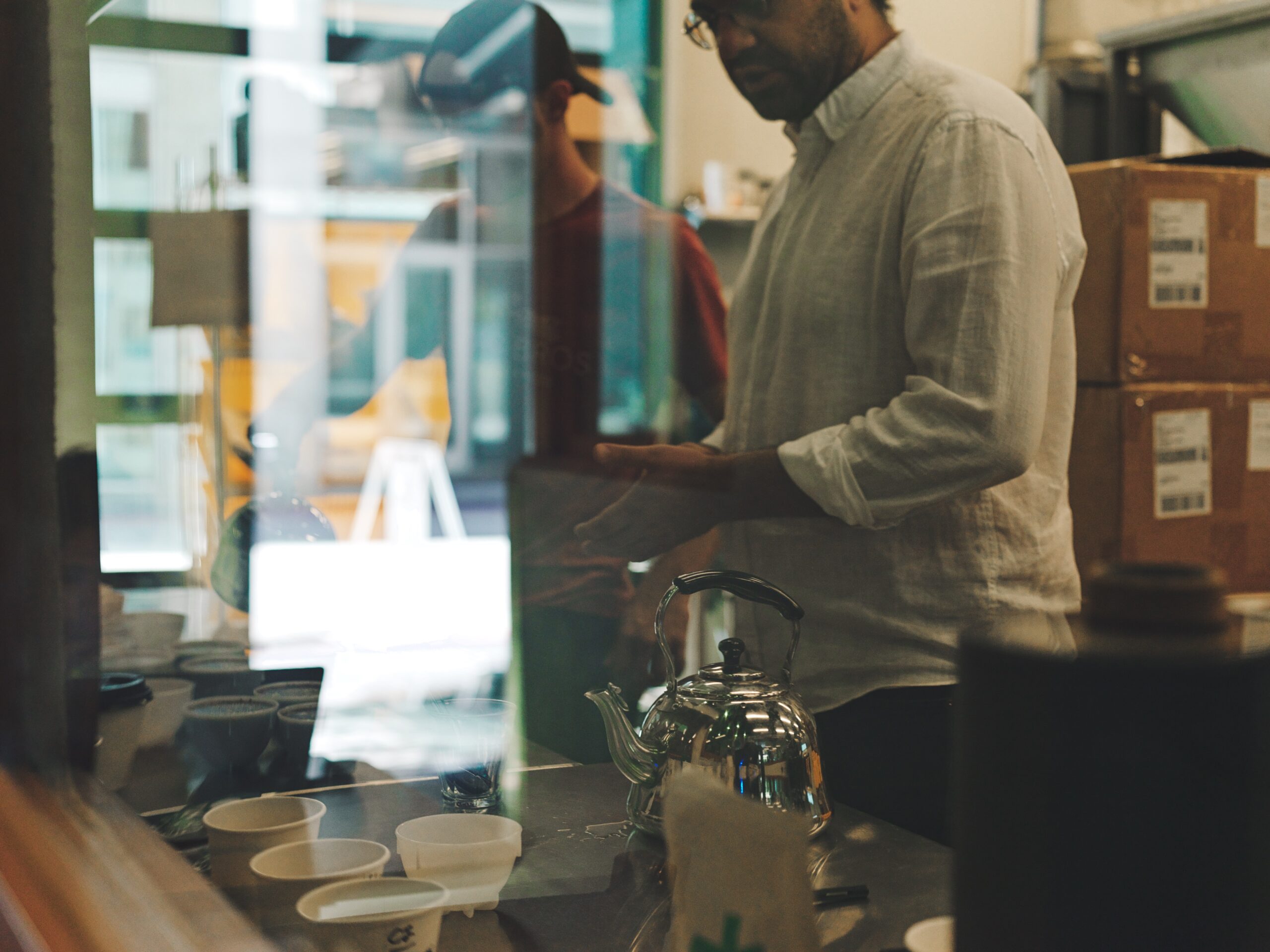 Two people prepare coffee behind a café counter with a metal kettle and tasting cups, viewed through a glass window with reflections.